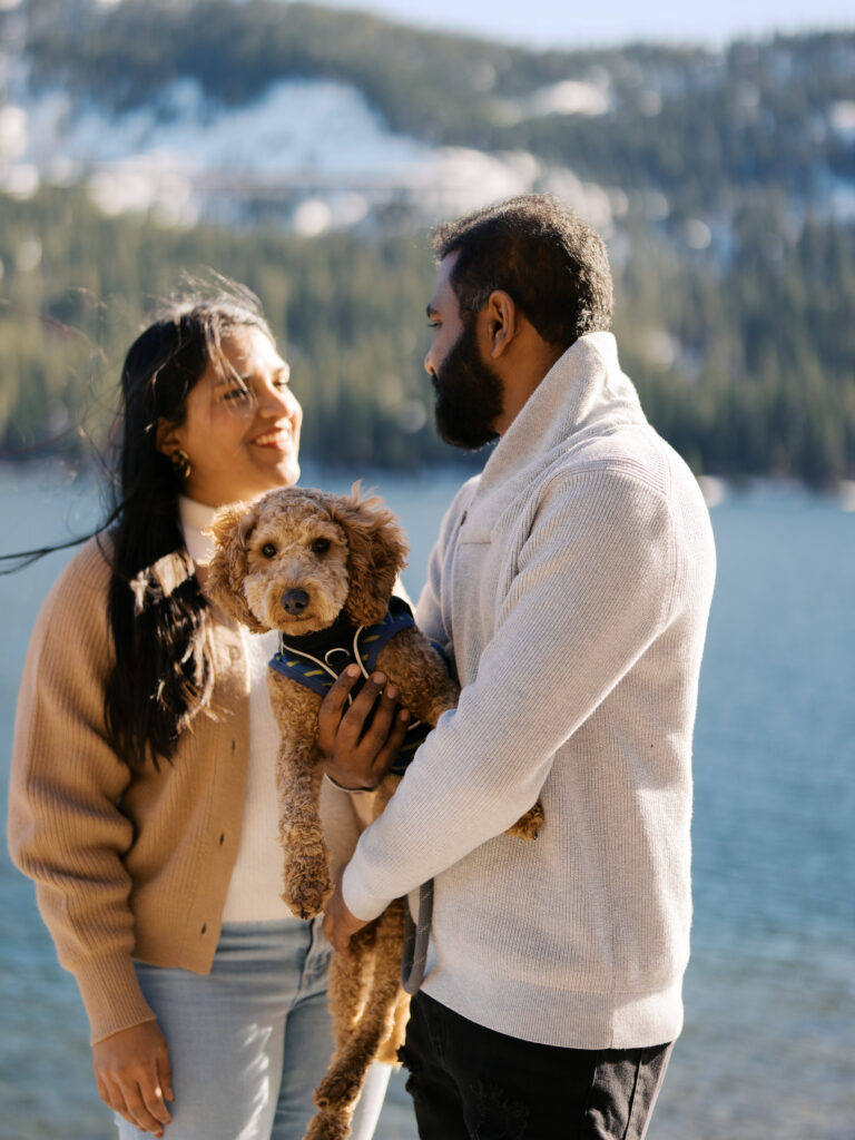 Couple smiling at each other while holding dog in their arms, the background shows donner lake and somre pie trees 