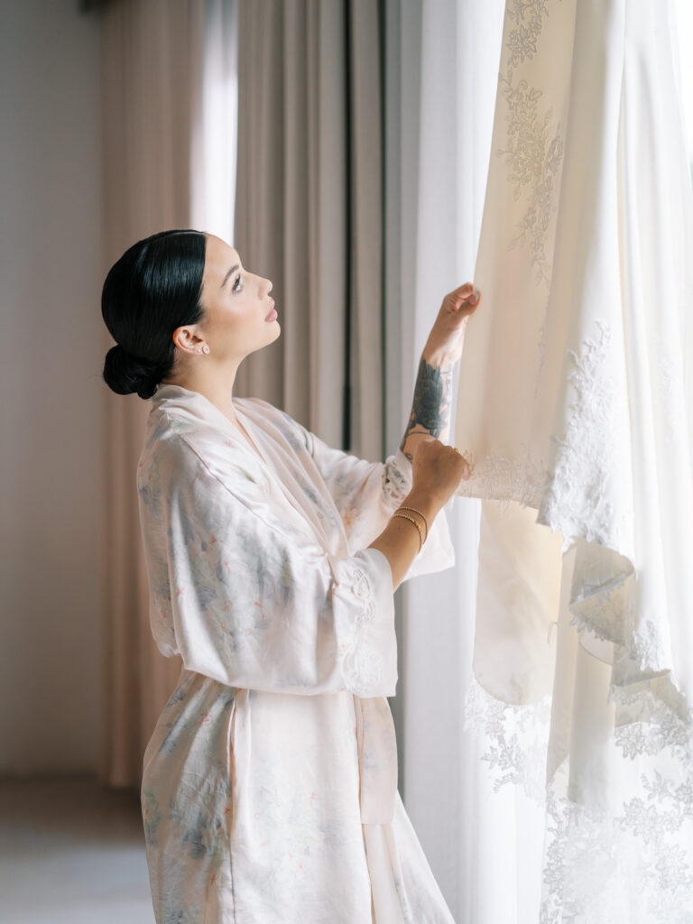 Bride in floral robe holding wedding dress up to window light, Tampa destination wedding photography