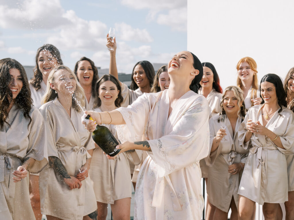 Bride popping champagne with bridesmaids in matching satin robes on rooftop, Tampa wedding getting ready