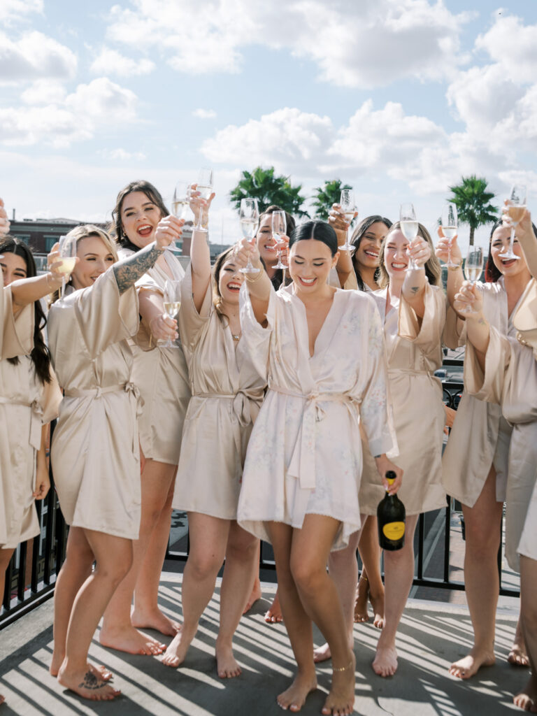 Bride and bridesmaids in champagne satin robes raising glasses on rooftop, Tampa wedding morning