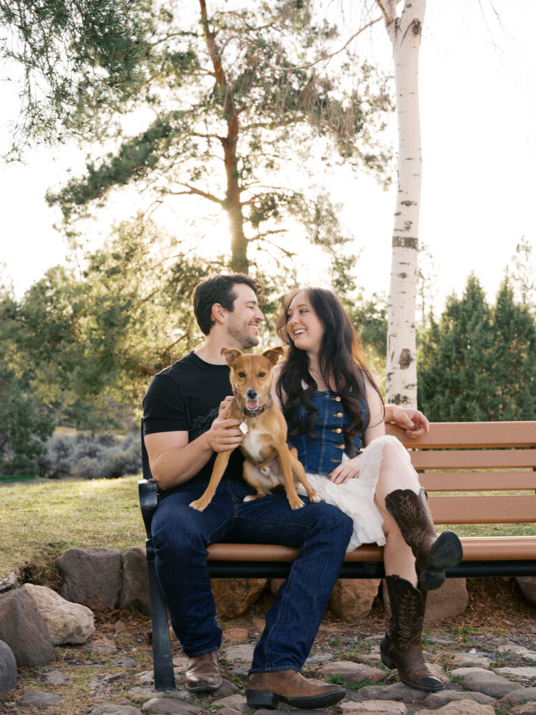 Couple sitting down with their dog in a bench smiling at each other captured by a Reno photographer in a couples session