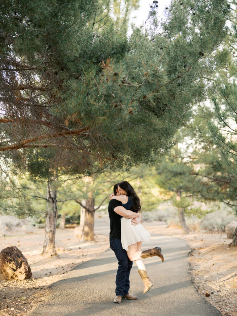 Boyfriend lifts and hugs girlfriend, she wears cowboy boots and a white skirts captured by a Reno photographer  in a couples session